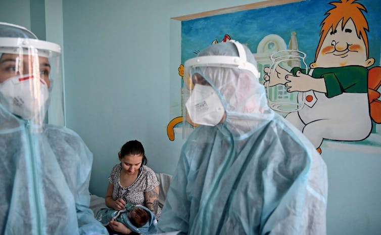 Une femme tient un masque à oxygène sur le visage de son enfant, derrière du personnel soignant en tenue de protection